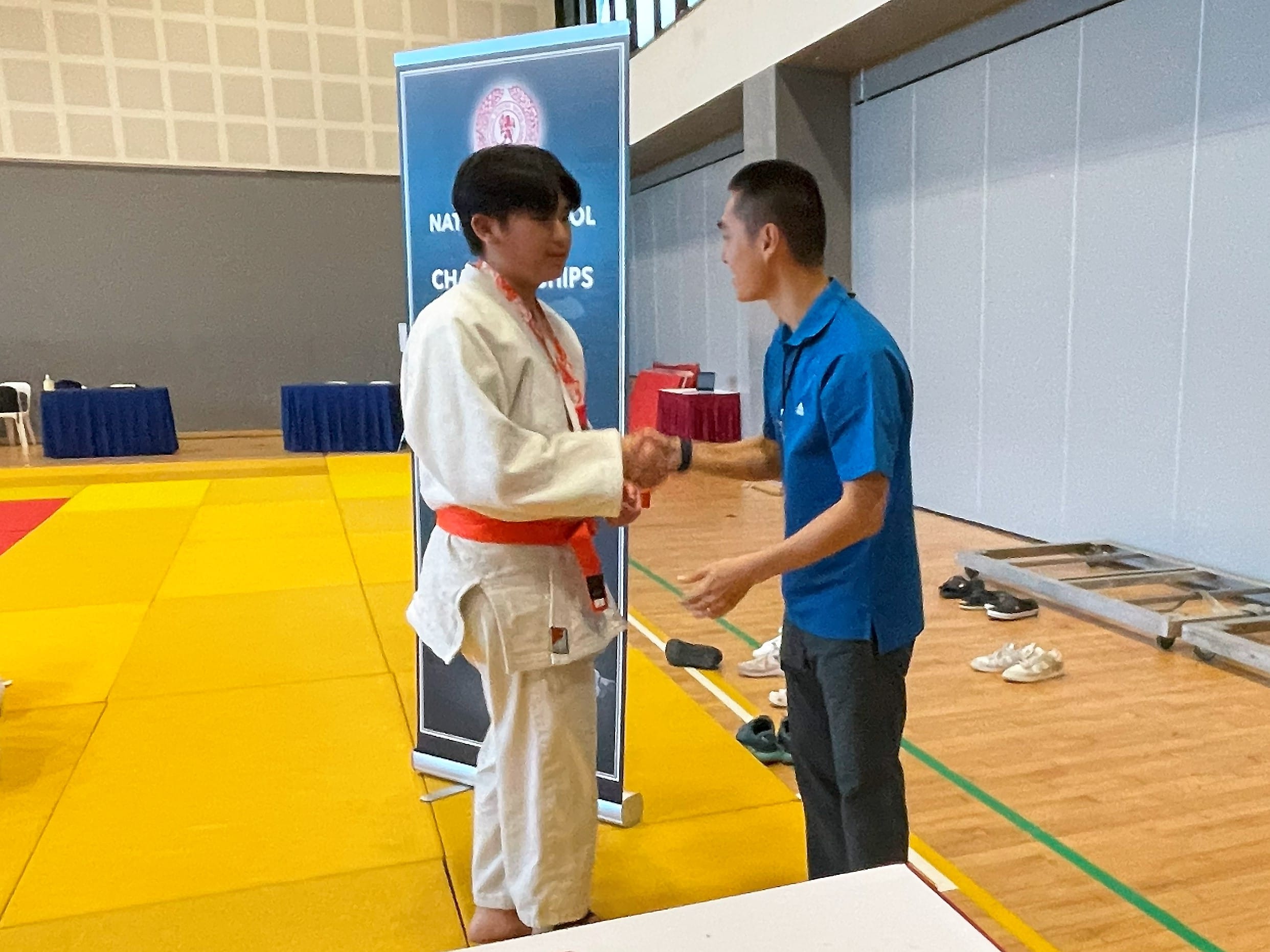 A judoka in a white gi shakes hands with an official beside a National School Games Championships Judo banner.